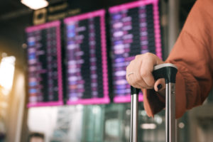 Close up of woman hand holding suitcase standing at the airport checking arrival departure board with the flight schedule in background.